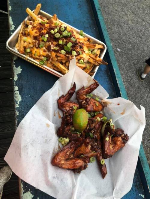 A tray of loaded cheese fries topped with bacon and green onions, alongside a serving of glazed chicken wings with lime.