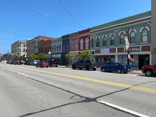 A sunny street view of a small town with colorful storefronts and parked cars along the road.
