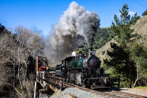 A vintage steam train crosses a bridge, releasing a plume of smoke against a backdrop of trees and hills.