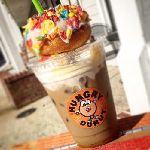 A colorful donut topped with cereal sits on a frosty drink from Hungry Donut, featuring a playful logo.