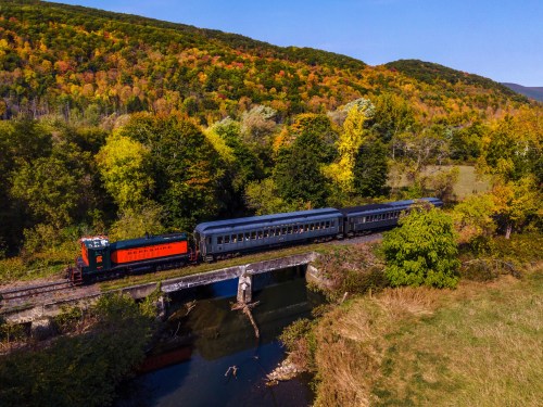 A colorful train travels over a bridge surrounded by autumn foliage and a serene landscape.