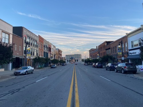 A wide street lined with shops and parked cars, under a clear blue sky with wispy clouds.