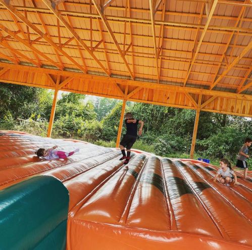 Children playing on a large inflatable bounce surface under a wooden shelter, surrounded by greenery.