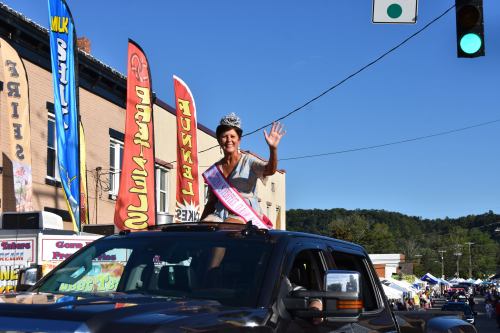 A woman wearing a crown and sash waves from the top of a car during a parade, with colorful banners in the background.