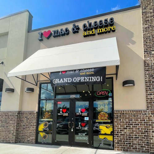 Exterior of "I Love Mac & Cheese and More" restaurant with a grand opening banner and open sign. Bright, inviting atmosphere.