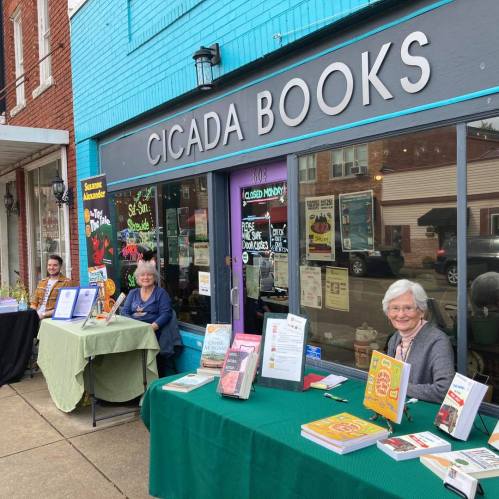 Two women and a man sit at tables outside Cicada Books, showcasing various books and promotional materials.