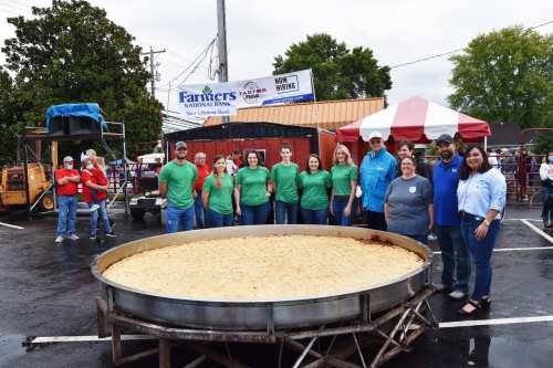 A group of people in green shirts stands around a large, round dish of food at an outdoor event.