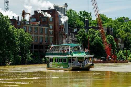 A green boat with passengers sails on a muddy river near an industrial area with smoke and cranes in the background.