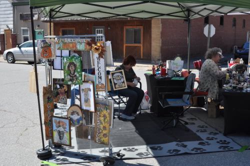A craft market scene with artwork displays, two vendors sitting under a tent, and a parked car in the background.