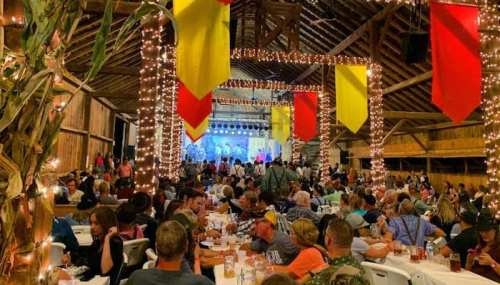 A lively barn filled with people dining and enjoying a performance, decorated with colorful banners and string lights.