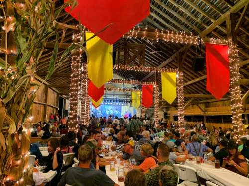 A lively barn event with people dining, colorful banners, and string lights, creating a festive atmosphere.