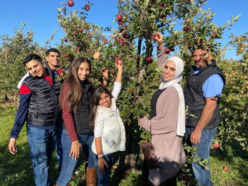 A family stands together in an apple orchard, picking apples from trees on a sunny day.