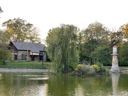 A serene pond scene featuring a log cabin, a lighthouse, and lush greenery under a clear sky.