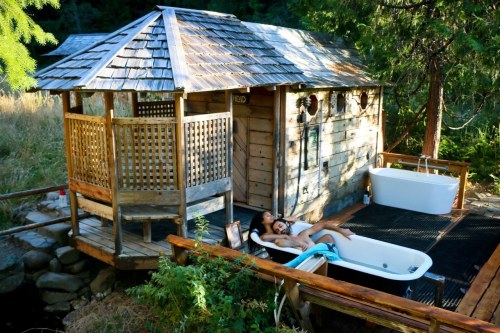 A cozy wooden cabin beside a hot tub, surrounded by greenery and a peaceful outdoor setting.