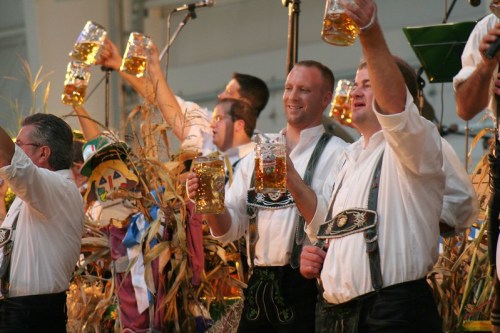 A group of men in traditional attire raise large beer mugs in celebration at a festive event.