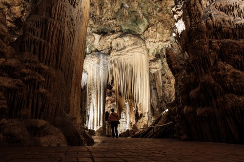 Two people explore a stunning cave with dramatic stalactites and stalagmites illuminated by soft lighting.
