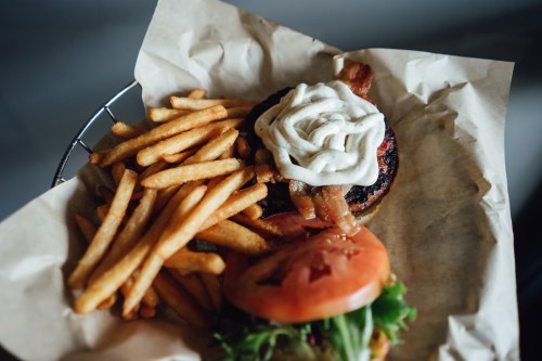 A burger with bacon, lettuce, tomato, and mayonnaise, served with a side of golden French fries on parchment paper.