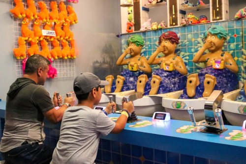 A man and a boy play a game at a colorful arcade, interacting with whimsical mermaid figures at sinks.