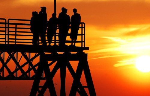 Silhouetted figures stand on a pier against a vibrant sunset, with warm colors illuminating the sky.