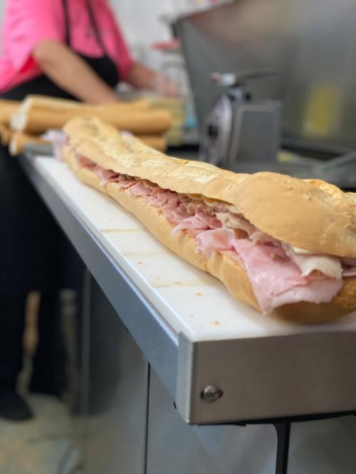 A long sub sandwich filled with layers of ham on a countertop, with a person in the background preparing food.
