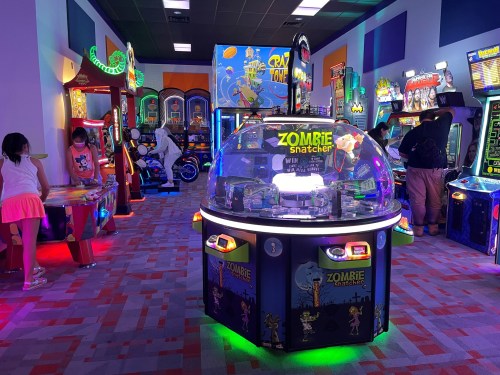 A vibrant arcade filled with colorful games and glowing lights, featuring a "Zombie Snatcher" game in the center.