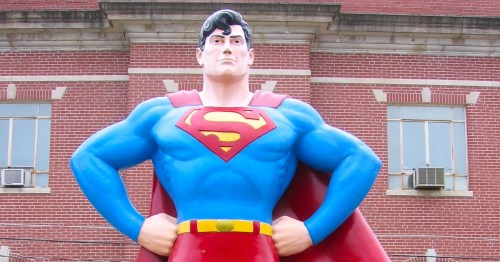 A large Superman statue stands confidently with hands on hips, wearing his iconic blue and red costume against a brick background.
