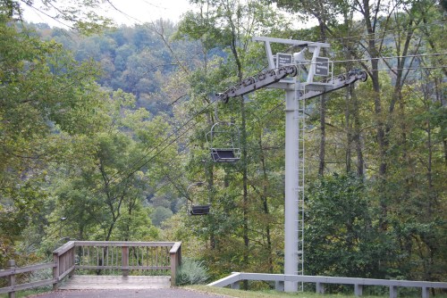 A chairlift stands among trees, with a scenic view of a green hillside in the background.