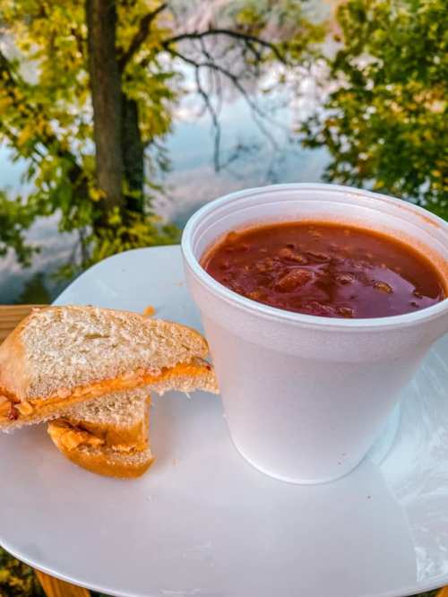 A cup of chili next to a half-eaten sandwich on a plate, with a scenic view of trees and water in the background.