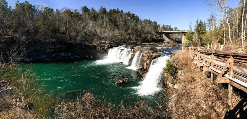A scenic view of a waterfall cascading into a turquoise river, surrounded by trees and a bridge in the background.