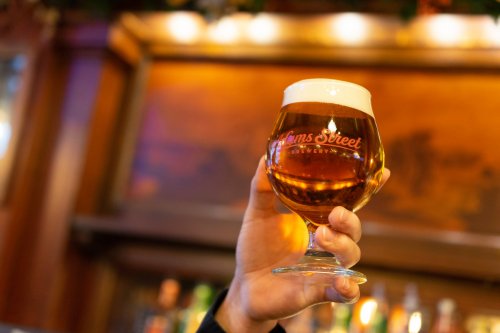 A hand holds a glass of amber beer with a frothy head, featuring the logo of Clams Street Brewery.