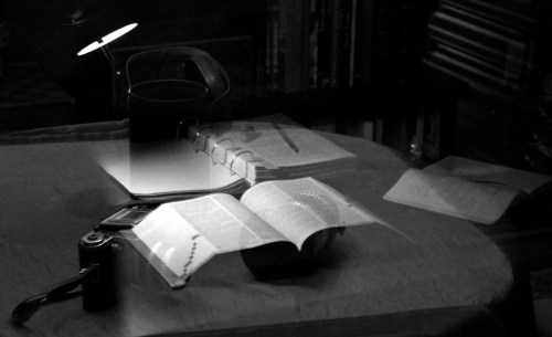 A dimly lit table with an open book, a camera, a notebook, and a lamp casting soft shadows.