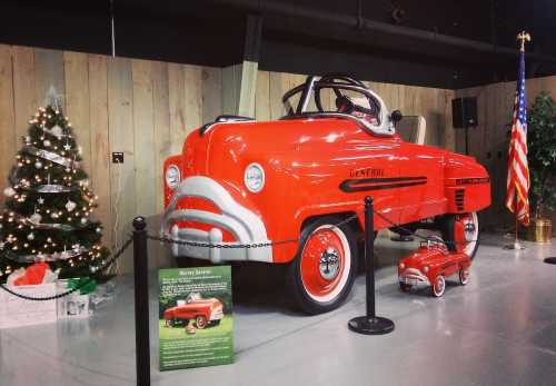 A vintage red car on display next to a Christmas tree, with a small matching toy car nearby.