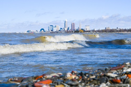 Waves gently lap at a rocky shore with a city skyline in the background under a clear blue sky.