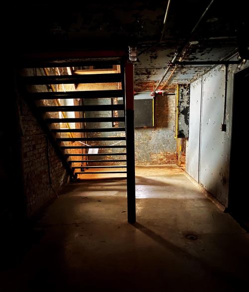 Dimly lit basement with a staircase, exposed brick walls, and a concrete floor. Shadows create a mysterious atmosphere.