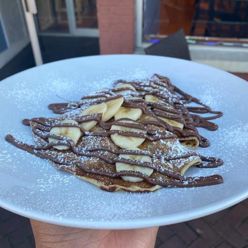 A plate of crepes topped with banana slices, drizzled with chocolate, and dusted with powdered sugar.