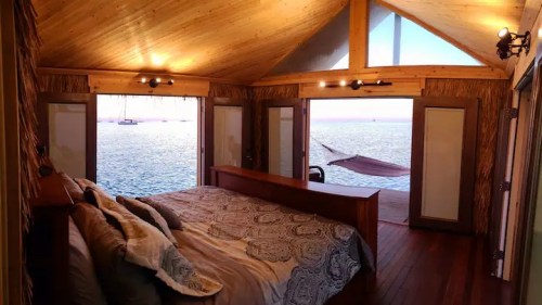 Cozy bedroom with wooden decor, large windows overlooking the ocean, and a hammock outside. Warm, inviting atmosphere.