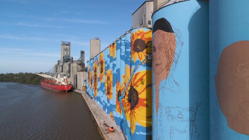 Colorful mural on grain silos by a river, featuring sunflowers and a portrait, with a cargo ship docked nearby.