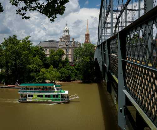 A boat glides on a river beneath a metal bridge, with historic buildings and a church steeple in the background.