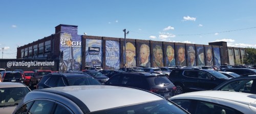 Mural of Van Gogh's portraits on a building, surrounded by parked cars under a clear blue sky.