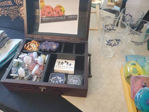 A wooden box labeled "Adventure Away" filled with game tokens, dice, and various colorful items on a tabletop.