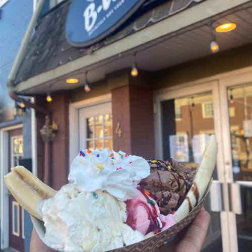 A hand holding a banana split with three scoops of ice cream, whipped cream, and sprinkles in front of a shop.