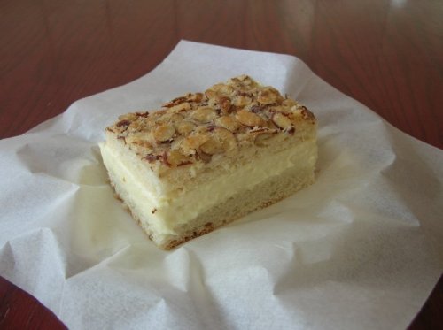 A square slice of layered dessert with a creamy filling, topped with sliced almonds, on a white paper napkin.