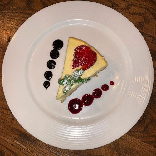 A slice of cheesecake topped with a strawberry, garnished with berry sauces on a white plate.