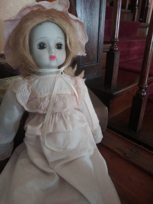 A vintage doll with blonde hair and a pink dress, sitting on a wooden surface near a staircase.