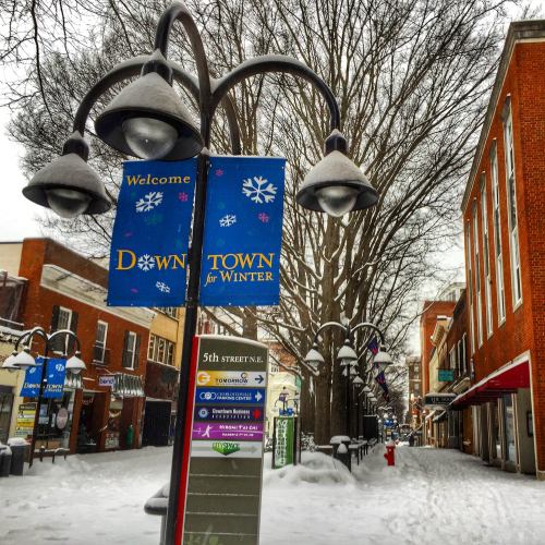 Snow-covered street in downtown with winter banners and lampposts, lined with shops and trees.