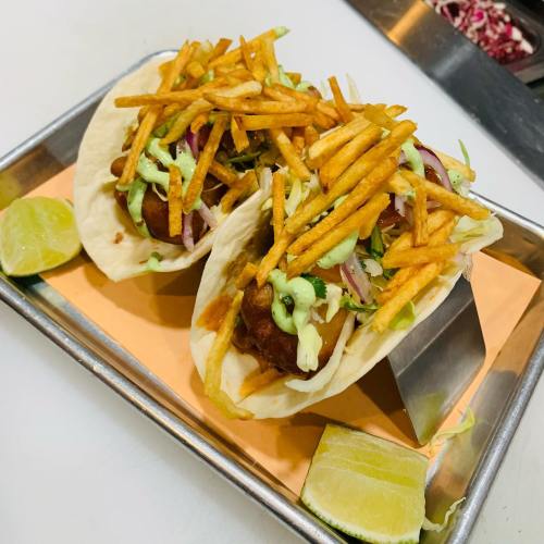 Two tacos filled with crispy toppings, drizzled with sauce, served on a tray with a lime wedge.