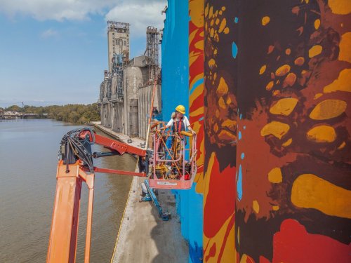 Workers on a lift painting a colorful mural on a large wall by a river, with industrial buildings in the background.