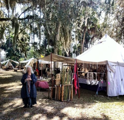 A woman in a long coat stands in a market with tents and wooden crafts, surrounded by trees and Spanish moss.