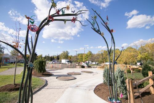Colorful metal sculptures arch over a winding path in a park, surrounded by trees and blue skies with fluffy clouds.