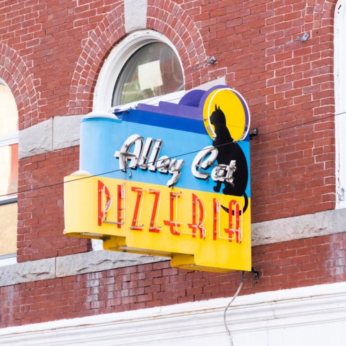 Colorful sign for "Alley Cat Pizzeria" featuring a cat silhouette against a blue background on a brick building.